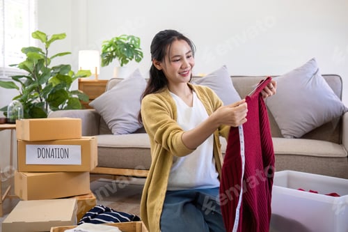 Preview: Cute Asian woman sitting next to sofa in living room sorting unwanted clothes for donation.