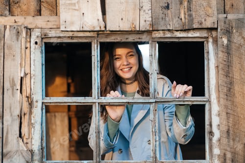 Preview: smiling girl looks out the window from an old wooden house, evening sunlight