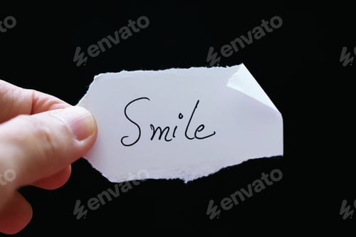 Preview: Hand holding white piece of paper with word “Smile” closeup background