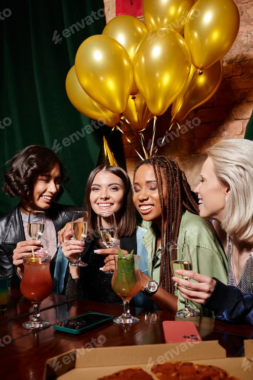 Preview: happy woman in party hat with champagne near multiethnic girlfriends and birthday balloons in bar