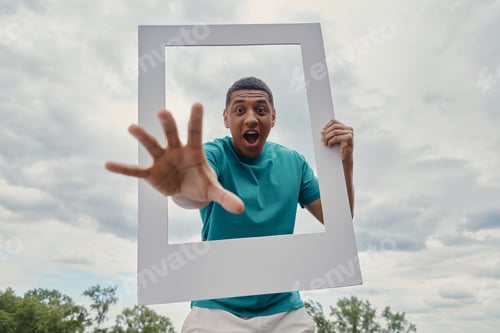 Preview: Playful mixed race man looking through a picture frame while standing outdoors
