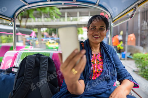 Preview: Mature Indian woman sitting in tuk tuk and using mobile phone