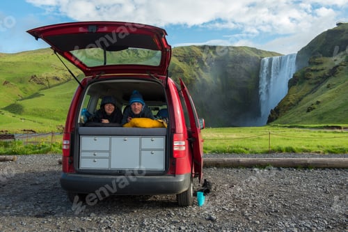 Preview: Couple relaxing in a red minivan with opened back door. Skogafoss waterfall, Iceland