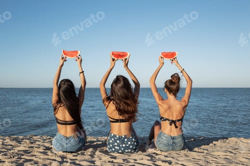 Preview: Back view of three dark-haired girls in jean shorts sitting on the sand near the sea holding