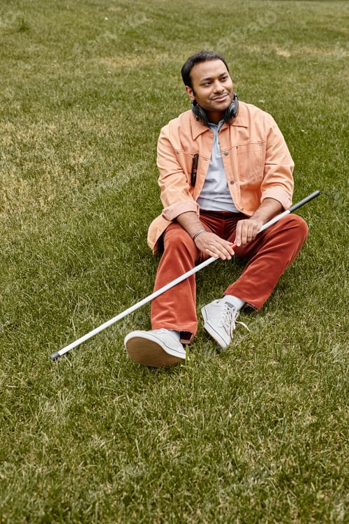Preview: joyful indian blind man in orange jacket sitting on grass with headphones and walking stick
