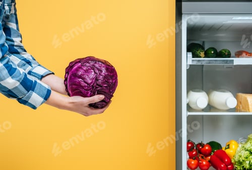 Preview: cropped view of woman holding red cabbage near open fridge with fresh food on shelves isolated on