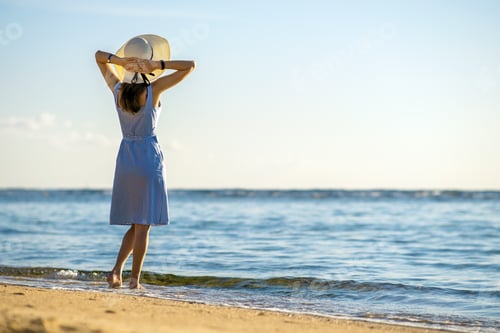 Preview: Young woman in straw hat and a dress standing alone on empty sand beach at sea shore