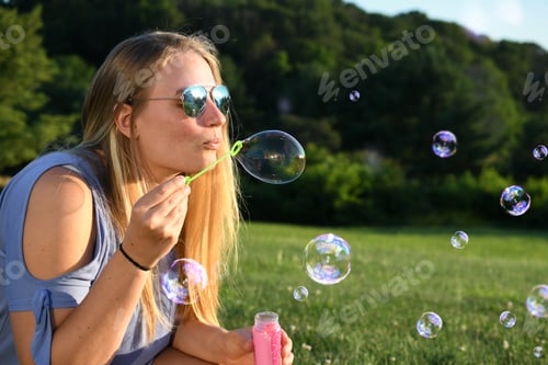Preview: Blonde woman with aviator sunglasses blowing bubbles at golden hour on a warm summer evening.