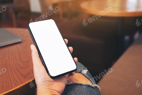 Preview: Hand holding black mobile phone with blank desktop screen with laptop computer on the table