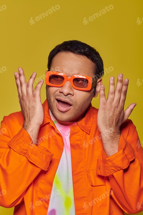 Preview: confused indian man in orange sunglasses looking away and gesturing on yellow background, expressive