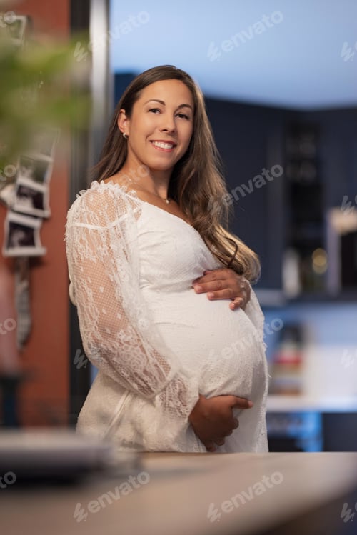 Preview: Pregnant woman smiling warmly in a kitchen setting wearing a white dress