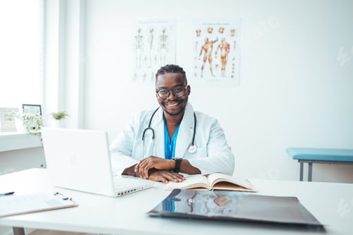 Preview: Portrait Of Smiling male Doctor Wearing White Coat With Stethoscope Sitting Behind Desk In Office.