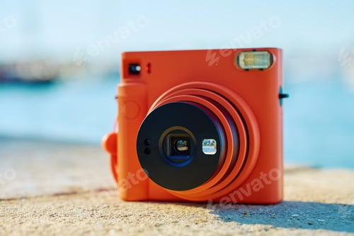 Preview: Orange instant camera on rock with seaside background