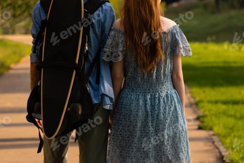 Preview: Crop unrecognizable couple walking along park path together with longboard backpack at sundown