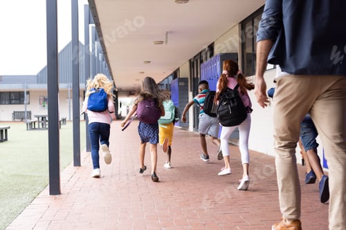 Preview: Rear view of diverse male teacher with children running in elementary school corridor, copy space