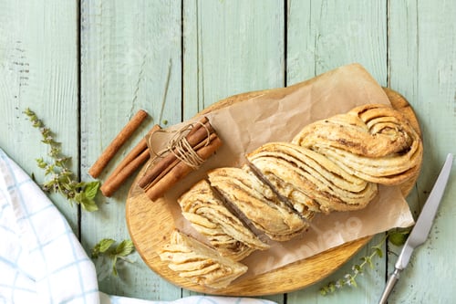 Preview: Braided cinnamon sweet bread on rustic wooden board with cinnamon sticks and herbs.
