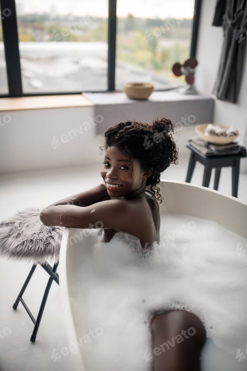 Preview: Beaming curly woman feeling cheerful while lying in foamy bath