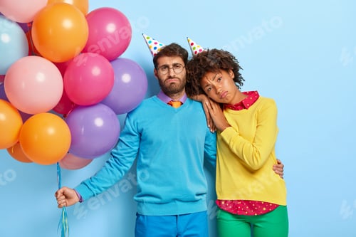 Preview: Tired upset diverse woman and man wear birthday hats, feel bored at party, dressed in colorful cloth
