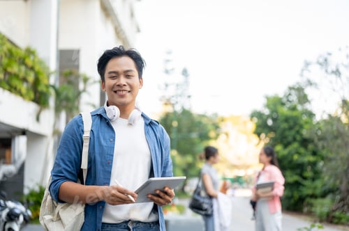 Preview: Smiling asian man student carrying a bag holding pen and tablet while standing in the university.