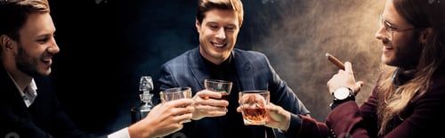 Preview: panoramic shot of happy men clinking glasses with whiskey on black with smoke