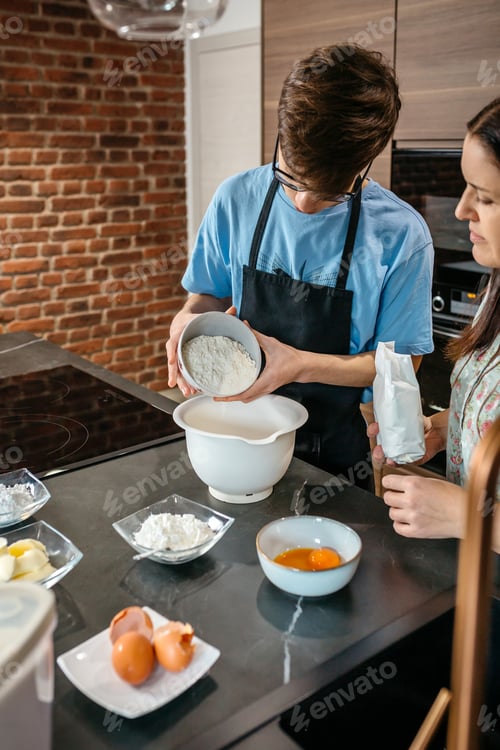 Preview: Teenager boy pouring flour into bowl while baking with mother in kitchen