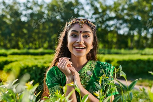 Preview: cheerful and pretty young indian woman in sari looking at camera near plants in park