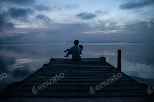 Preview: Silhouette of a father holding his child in his arms as dusk falls. They are sitting on a pier.