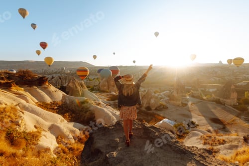 Preview: Happy girl in hat stands on mountain with flying air balloons on background. View from the back.