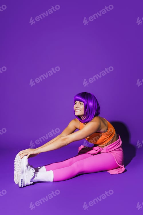 Preview: cheerful woman wearing colorful sportswear during stretching workout against purple background