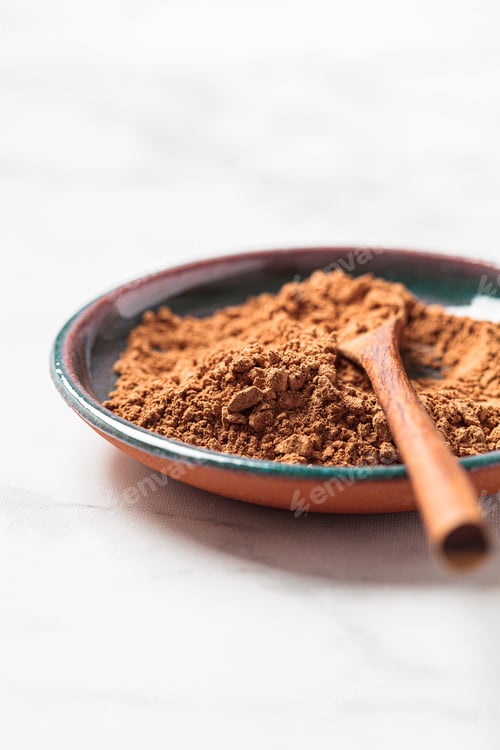 Preview: Raw organic cocoa powder on saucer with wooden spoon, white marble background. Super food concept.