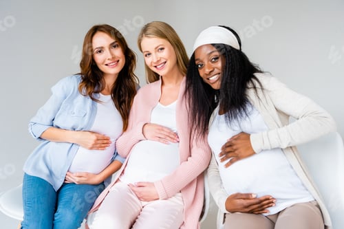 Preview: multicultural pregnant women sitting close together, touching bellies isolated on grey
