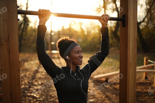 Preview: Happy African American female athlete doing chin-ups on exercise equipment at sunset.