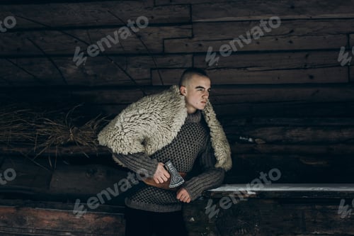 Preview: Ancient viking warrior with steel axe posing near wooden hut in scandinavian forest