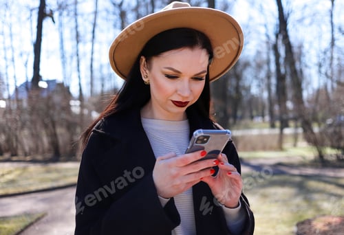 Preview: Beautiful young brunette girl with long hair in a hat using a mobile phone in the park