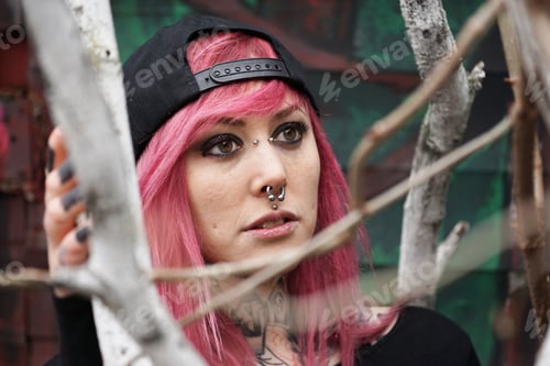 Preview: young woman with pierced face and tattooed neck behind tree branches