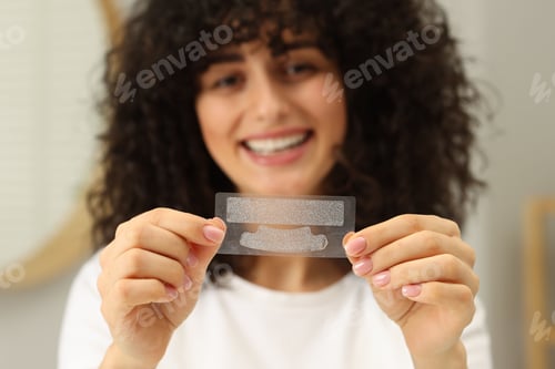 Preview: Young woman holding teeth whitening strips indoors, selective focus