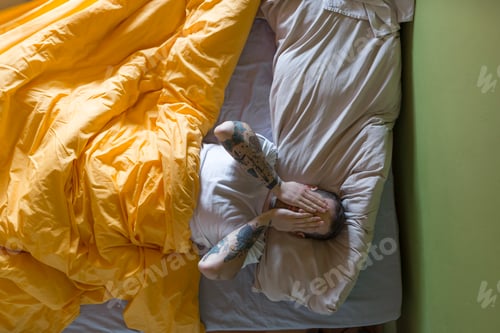 Preview: Tattooed man lying in bed, hands on eyes