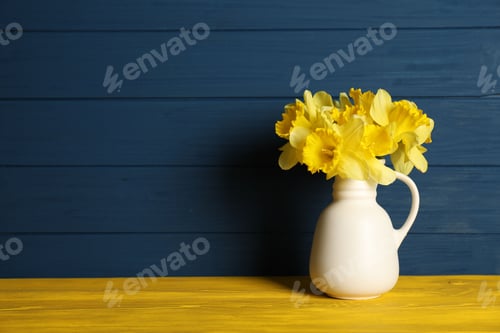 Preview: Beautiful daffodils in vase on yellow wooden table against blue background. Space for text