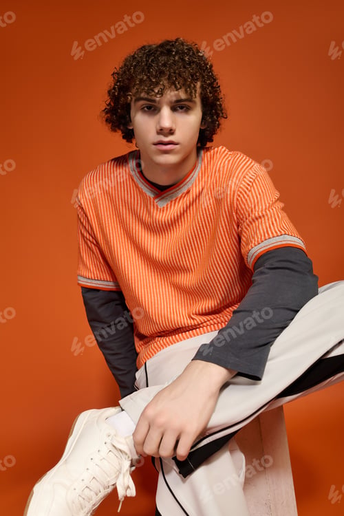 Preview: Curly-haired teenager in trendy casual wear against a vibrant orange backdrop