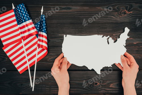 Preview: top view of arranged american flags and female hands with piece of blank map on wooden tabletop,
