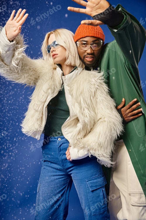 Preview: african american man and stylish woman posing in winter attire, gesturing under falling snow on blue