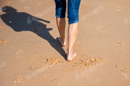 Preview: woman walking barefoot on beach during a sunny day, rear view, closeup