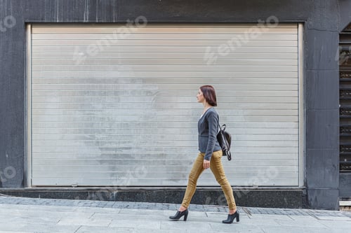 Preview: Woman walking in urban setting near a closed garage door in modern attire