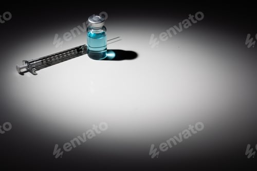 Preview: Syringe Needle and Medicine Vial With Light Blue Chemical Spot Lit on Reflective Background