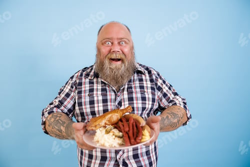 Preview: Jouful man with overweight holds plate of rich food standing on light blue background