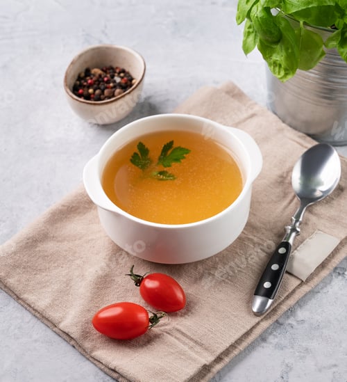 Preview: Meat broth with parsley in a white bowl on a light background with fresh herb, tomato and spoon