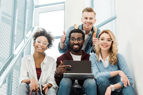 Preview: group of smiling multicultural students with laptop showing thumbs up in university