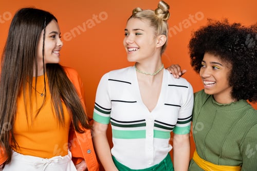 Preview: Portrait of positive and multiethnic teenage girlfriends in trendy outfits with makeup talking and