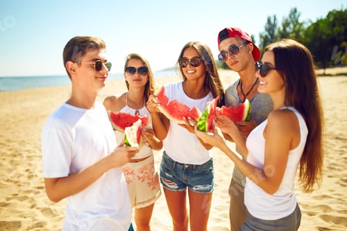 Preview: Group of friends having fun eating watermelon on beach. Excellent sunny weather. Beautiful figures.