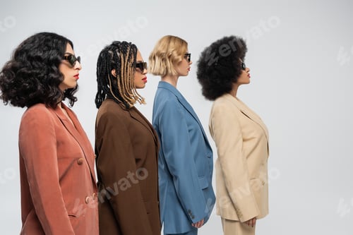 Preview: side view of stylish multiracial girlfriends in dark sunglasses and multicolored suits on grey